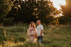 Pregnant couple standing in a sunlit meadow at golden hour, woman in pink dress, man in white shirt and jeans.