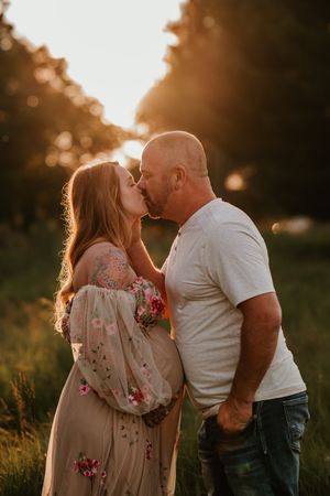 Couple sharing a tender kiss during golden hour sunset in a lush green field, woman in floral maternity dress.