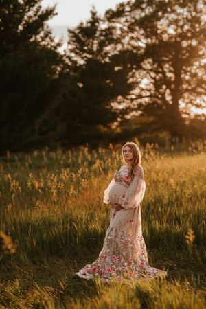 Pregnant woman in floral gown standing in golden field at sunset.