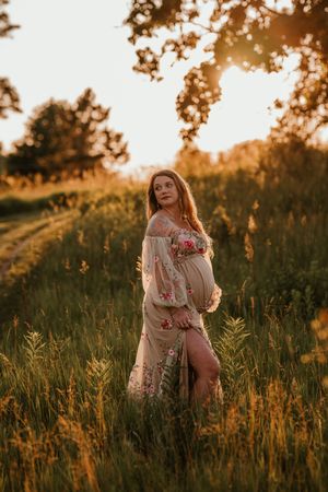 Pregnant woman in floral dress posing in tall grass during golden hour sunset.