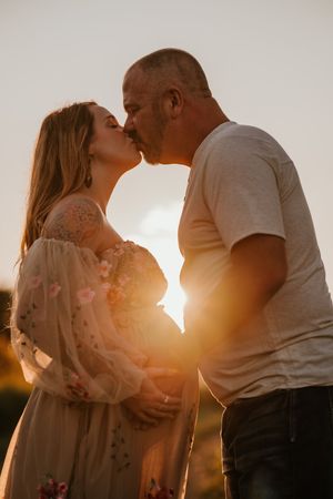 Couple sharing a kiss at golden sunset, pregnant woman in floral dress with partner in white t-shirt outdoors.