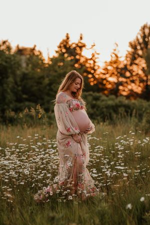 Pregnant woman in floral dress cradling baby bump in wildflower field at golden hour sunset.
