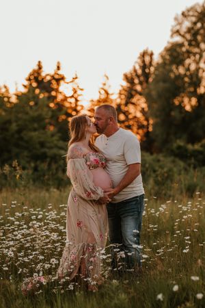 Couple sharing a kiss in a wildflower field at golden hour, woman in floral maternity gown with visible baby bump.