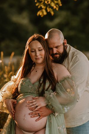 Couple sharing a tender maternity photo outdoors at golden hour, man embracing pregnant woman in green floral dress.