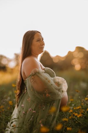 Pregnant woman in sheer green floral dress standing in golden field at sunset, cradling baby bump.