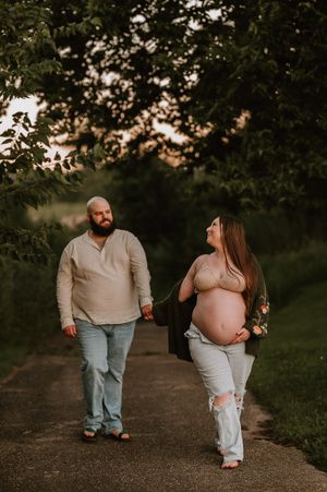 Pregnant couple walking hand-in-hand on a tree-lined path at golden hour, woman cradling baby bump.