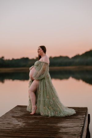Pregnant woman in green floral tulle gown standing on wooden dock at sunset by calm lake.