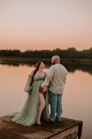 Expecting couple in green tulle maternity gown embracing on dock at golden hour by serene lake.