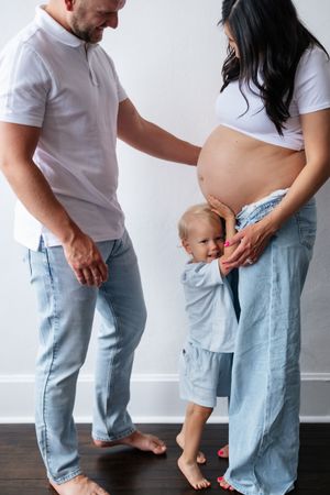 Happy family with pregnant mother, father, and toddler child posing together in casual denim outfits at home.