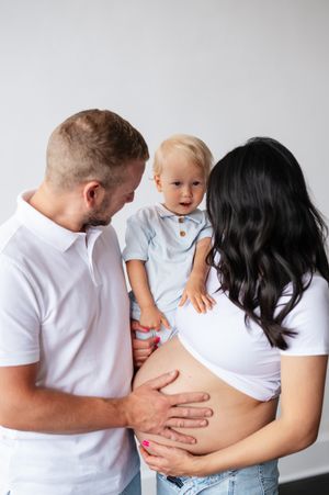 Pregnant woman and man holding toddler boy while touching baby bump during family maternity photo session.