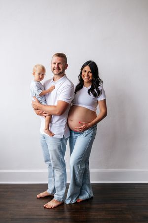 A happy family maternity photo with a pregnant woman, man, and toddler dressed in white and jeans.