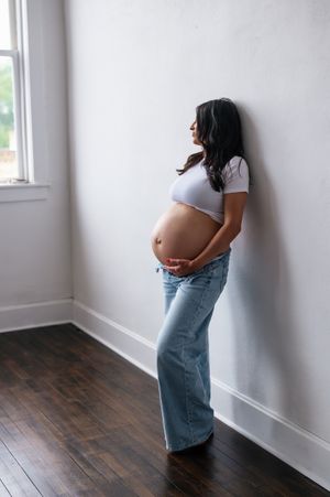 Pregnant woman in white crop top and jeans leaning against wall, cradling baby bump near window.