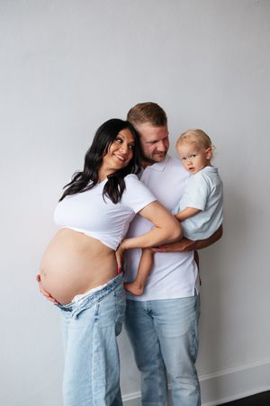 Pregnant woman in white crop top and jeans poses with partner holding toddler for maternity family photo.