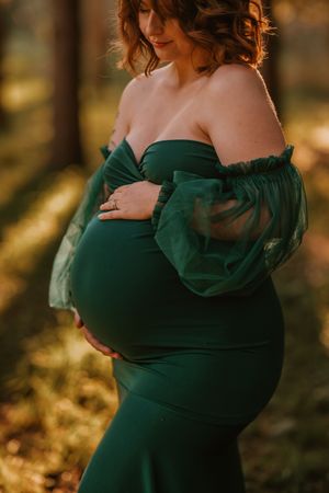 Pregnant woman in elegant emerald green off-shoulder gown with sheer sleeves, cradling her baby bump in a sunlit forest.