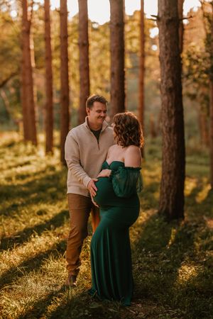 Couple sharing a tender moment in a sunlit pine forest, woman in green maternity gown held by man in beige sweater.