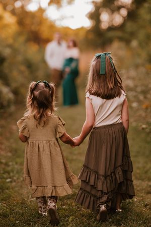 Two young sisters in boho dresses hold hands walking toward their parents in a golden autumn field.
