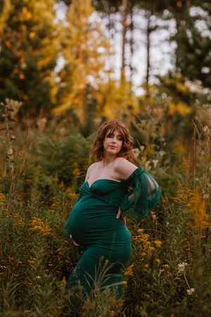 Pregnant woman in elegant green off-shoulder gown poses in a wildflower meadow during fall season.