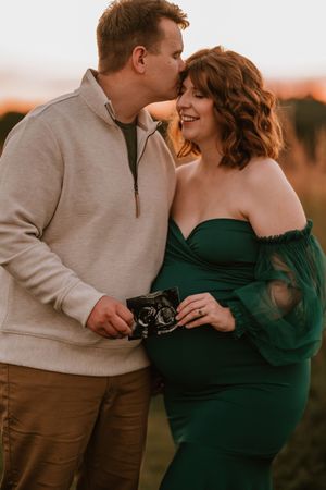 Couple holding ultrasound photos at golden hour, woman in green off-shoulder dress, man kissing her head.