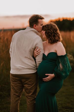 Couple sharing a tender moment at sunset in a field, pregnant woman in emerald green gown cradling her baby bump.