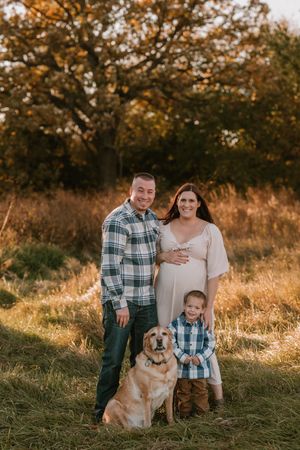 Family maternity photo with couple, young boy, and golden retriever in autumn field at golden hour.