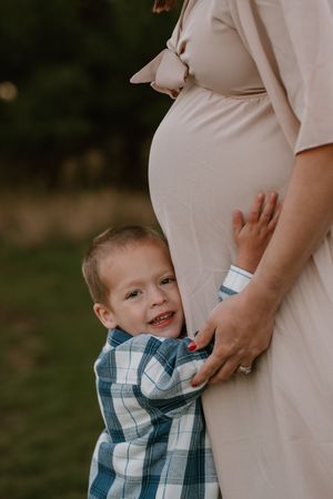 A young boy smiles while hugging his pregnant mother's belly during an outdoor maternity photo session.