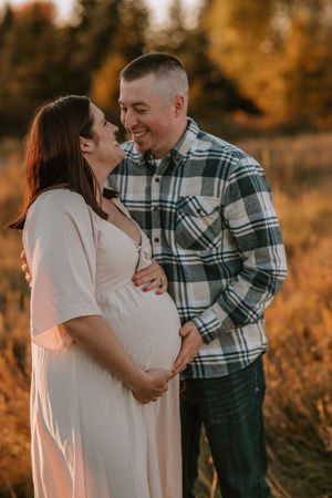 A couple smiles at each other outdoors at golden hour, the woman in a white dress showing her pregnant belly.