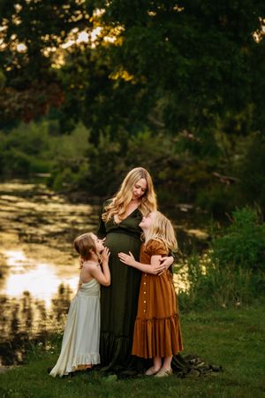 Pregnant woman in green dress with two daughters touching her belly near a sunlit pond.