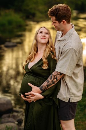 Pregnant woman in green dress with partner outdoors by a pond during golden hour maternity photo session.