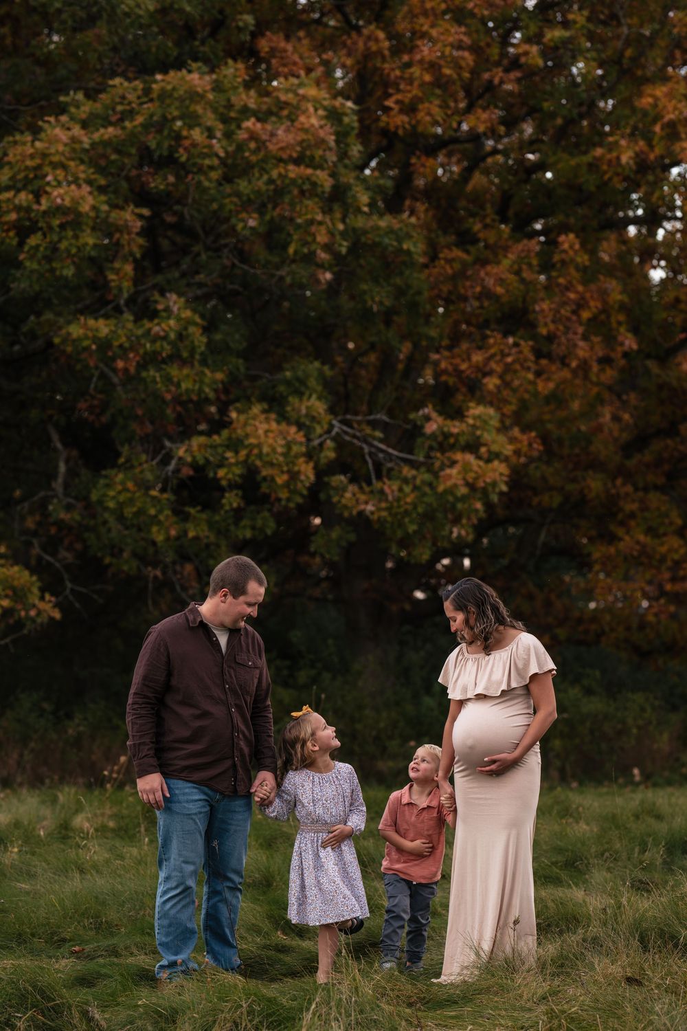 Pregnant woman in cream gown with family in autumn field, two children and husband smiling during maternity photo session.