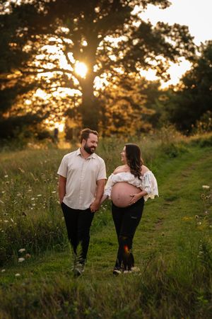 Pregnant couple walking through a sunlit meadow at golden hour, sharing a tender glance as woman cradles her baby bump.