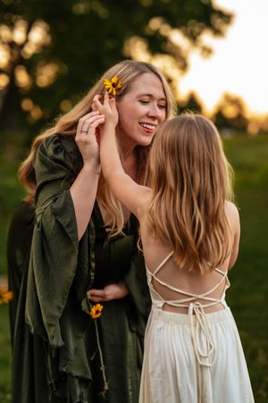 A smiling woman in a green dress has a sunflower tucked in her hair by a young girl in a white backless dress outdoors.