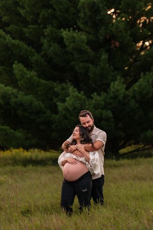 Couple embracing in a green meadow during a golden hour maternity photo shoot, man holding pregnant woman from behind.