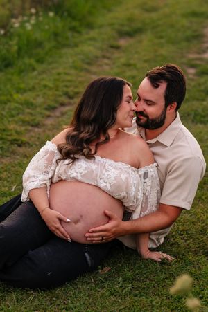 Couple sharing a tender moment outdoors, woman in lace maternity top cradling baby bump while partner embraces her on grass.