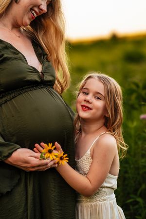 A young girl holds yellow flowers beside her pregnant mother in a green dress during a golden hour outdoor photoshoot.