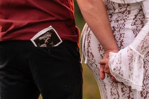 Pregnant couple holding hands, man with ultrasound photo in pocket, woman in white lace dress, pregnancy announcement photo.