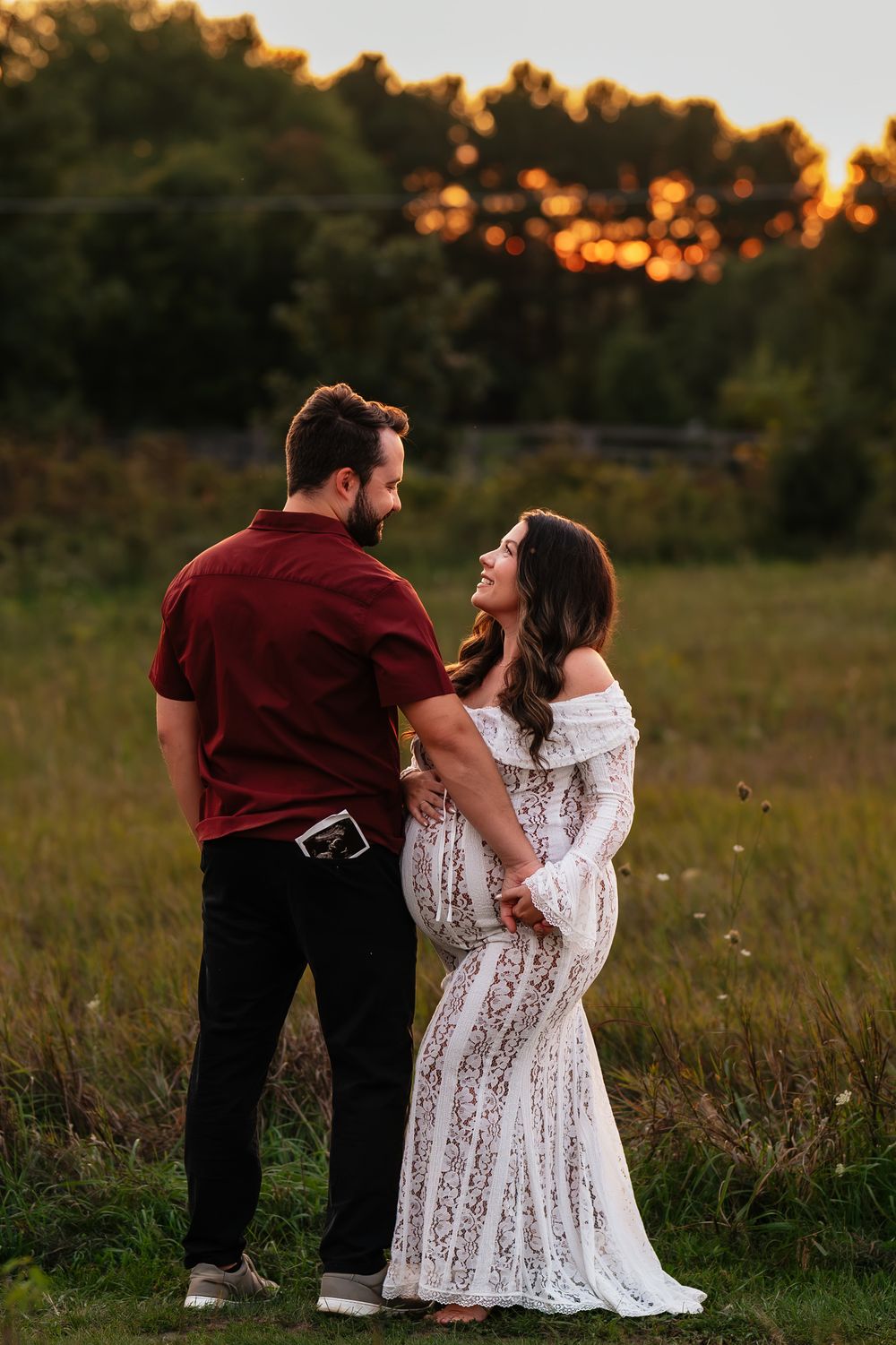 Couple sharing a tender moment at golden hour in a field, woman in white lace maternity gown holding ultrasound photo.