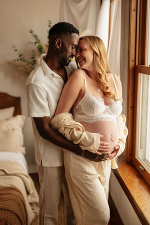 Interracial couple sharing tender moment, pregnant woman in white lace lingerie embraced by partner near sunlit window.