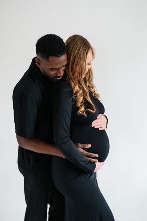 Interracial couple in matching black outfits, man tenderly embracing pregnant woman against white background.