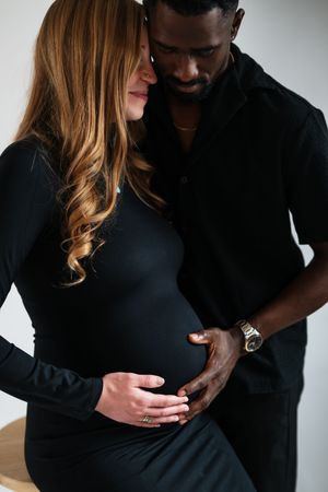 Interracial couple in black outfits tenderly cradling pregnant belly together in a studio setting.