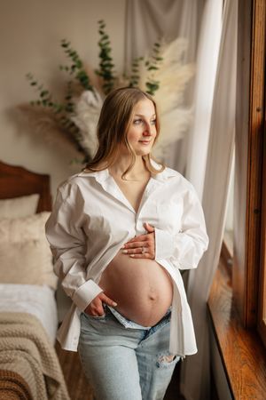 Pregnant woman in white shirt and jeans cradling baby bump near window in cozy bedroom.