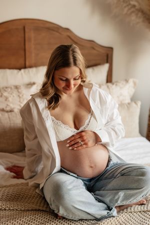 Pregnant woman sitting cross-legged on bed in white shirt and lace bralette, cradling her baby bump.