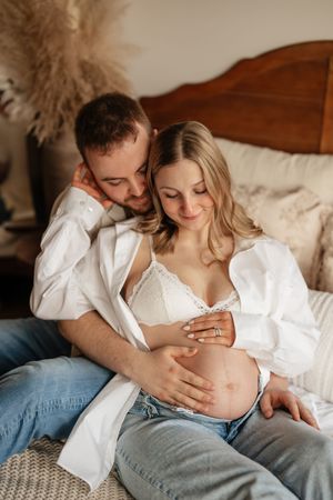 Couple sitting on bed, pregnant woman in white bra and open shirt, partner embracing her from behind, both touching baby bump.