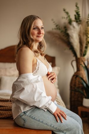 Smiling pregnant woman in white lace bra and open shirt sitting on bed, cradling baby bump in cozy bedroom.