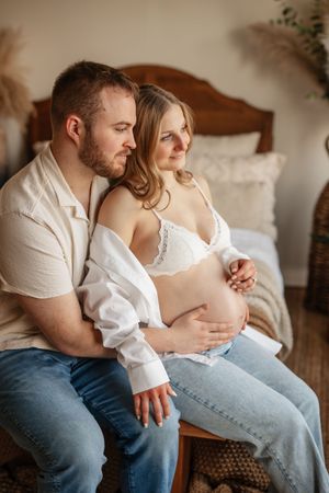 Couple sitting together on bench, man tenderly holding pregnant woman's belly in cozy bedroom setting.