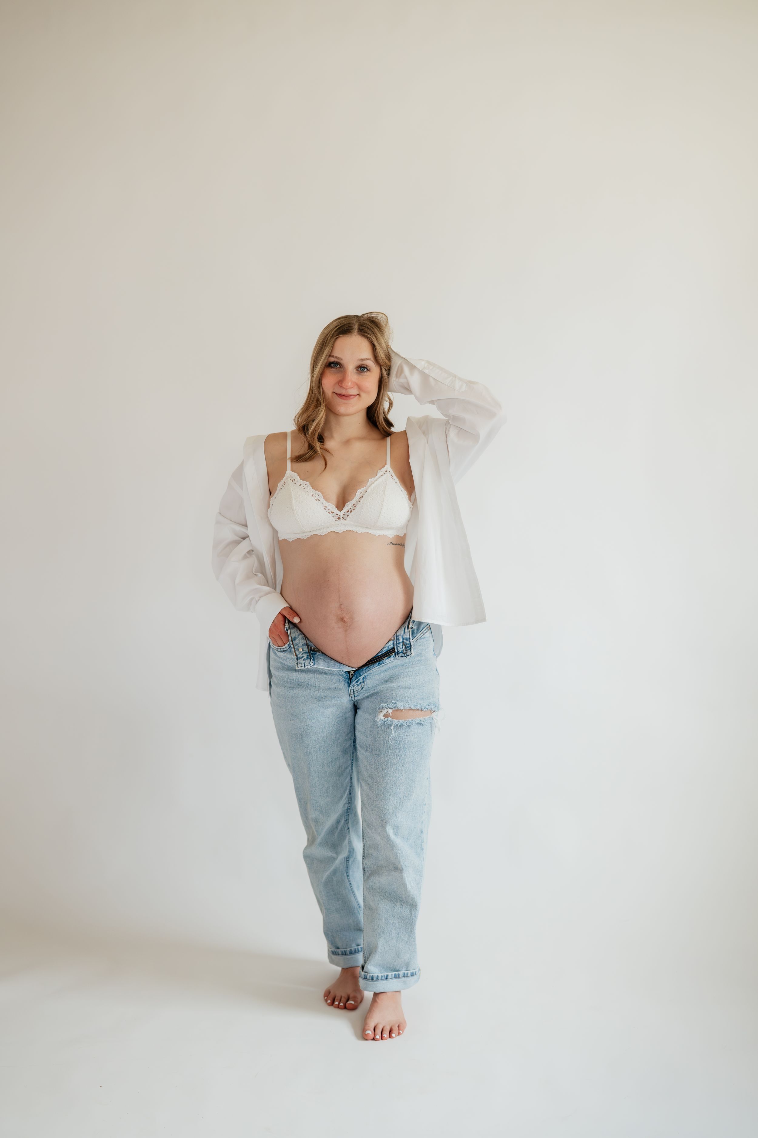 Pregnant woman in white lace bralette, open shirt, and distressed jeans posing against a white studio background.