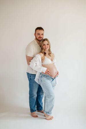 Couple maternity photo, man embracing pregnant woman in white bra and jeans, studio white background.