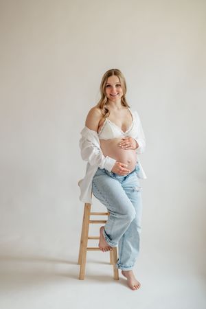 Smiling pregnant woman in white bra and jeans sits on wooden stool, cradling baby bump, wearing open white shirt.