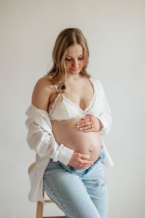 Pregnant woman in white lace bra and jeans cradling baby bump, wearing open white shirt, studio maternity photo.