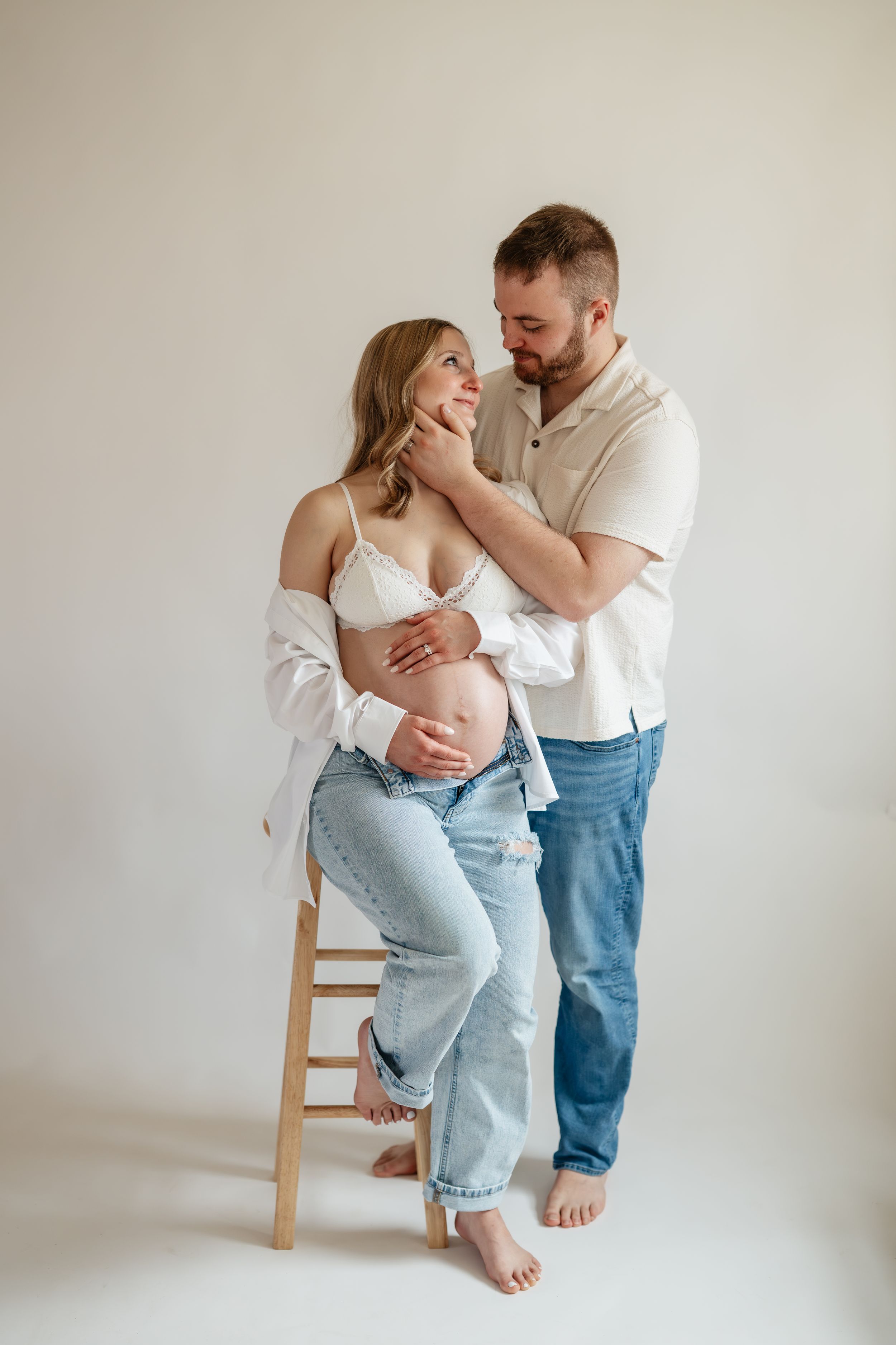 Pregnant couple in white studio, man tenderly touches woman's face as she sits on stool cradling baby bump.