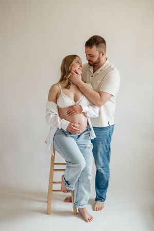 Pregnant couple in white studio, man tenderly touches woman's face as she sits on stool cradling baby bump.